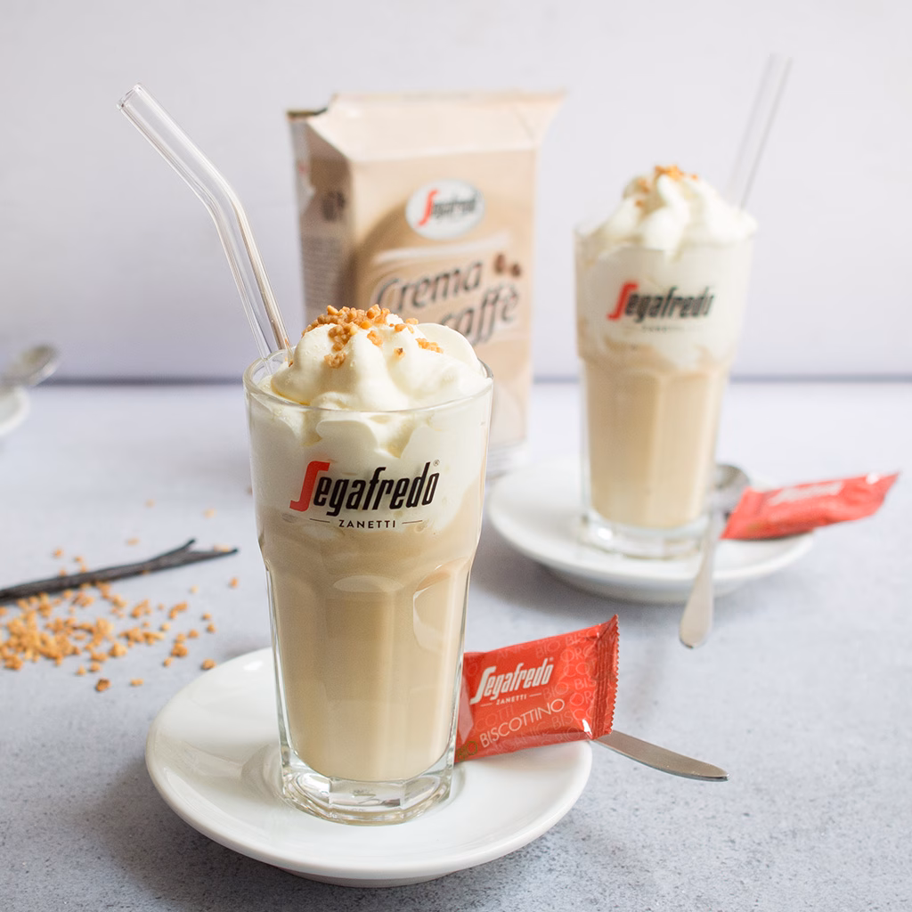 Imagebild Latte Macchiato-Glas mit Kaffeeeis und Schlagobers und einer Keksverpackung, im Hintergrund unscharf ein weiteres Glas und die Produktverpackung