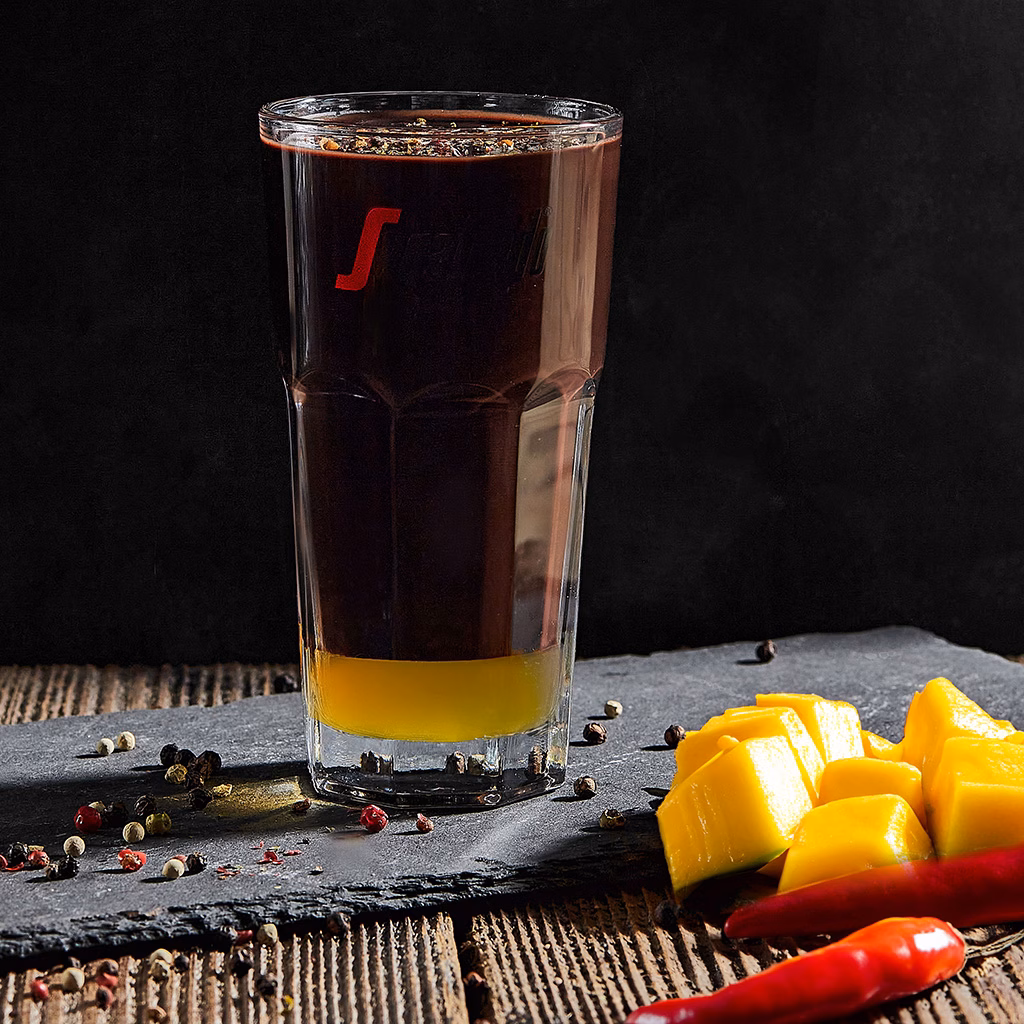 Imagebild Glas mit gelbem Mango-Sirup und Schokoladenpudding auf einer Schieferplatte, Mango und Chili im Vordergrund, dunkler Hintergrund