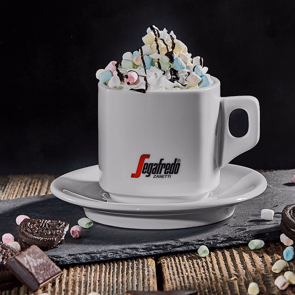 Imagebild weiße Tasse mit Schlagobers, kleinen Marshmallows und Schokoladensauce auf einer Schieferplatte, kleine Marshmallows und Kekse im Vordergrund, dunkler Hintergrund