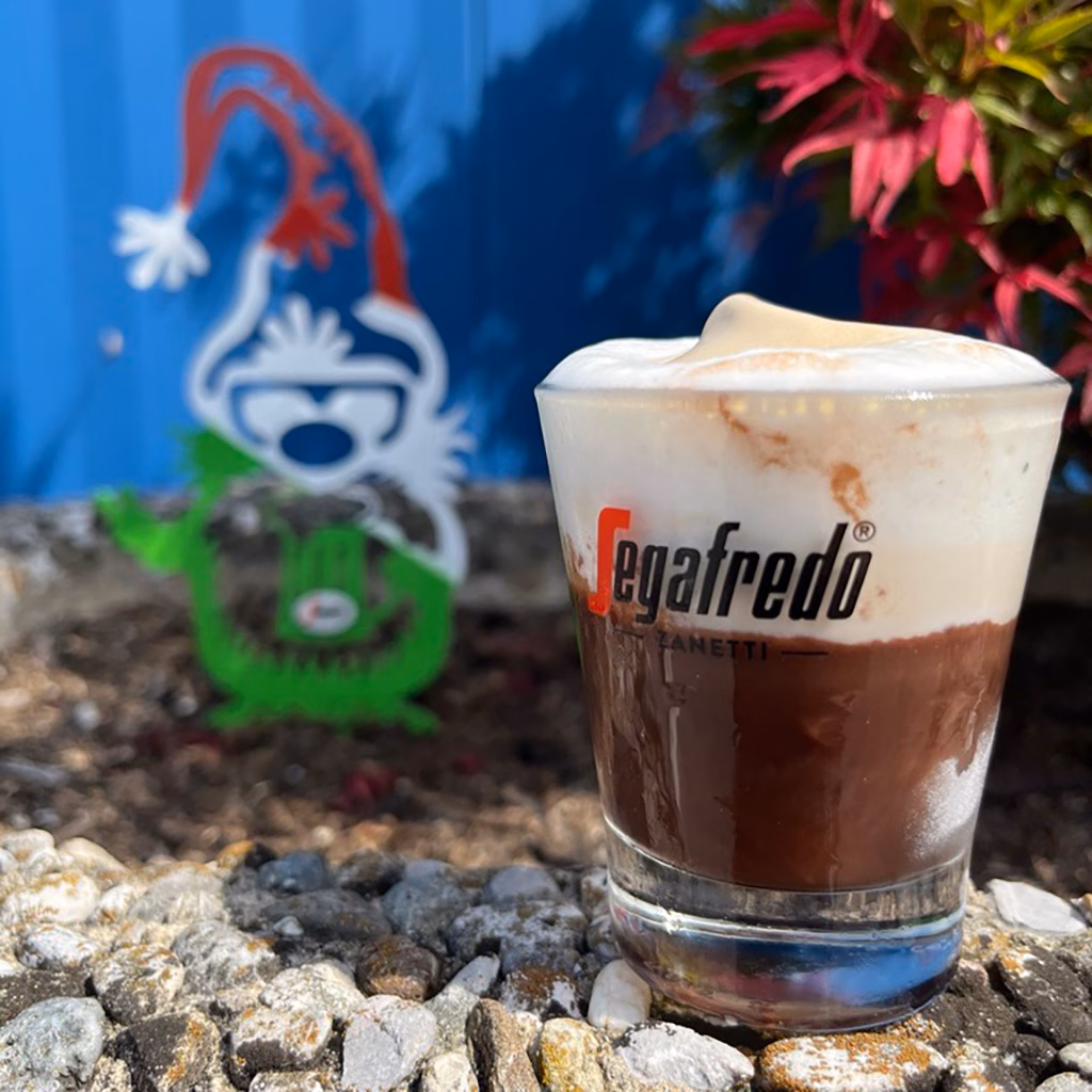 Imagebild Glas mit Schokoladenpudding, Milchschaum und halbgefrorenem Kaffeeeis, im Hintergrund eine bunte Metallfigur